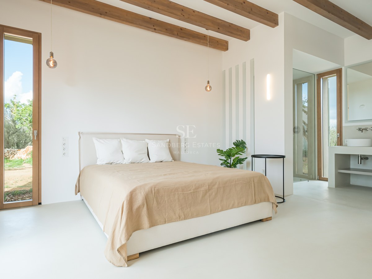 Minimalist bedroom featuring exposed wooden ceiling beams, white microcement floors, and en-suite bathroom.