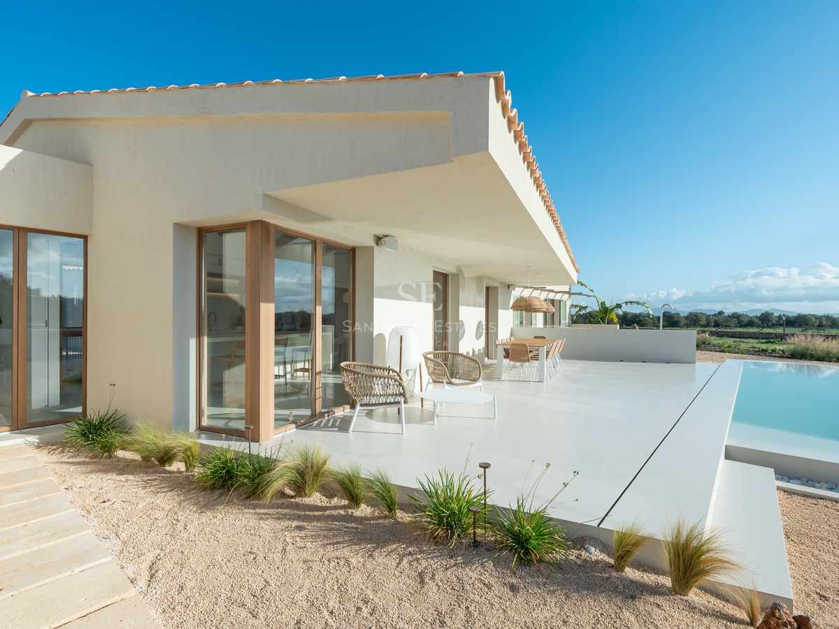 Modern villa terrace with white lounge furniture, wooden framed glass doors, and a turquoise infinity pool under clear sky.