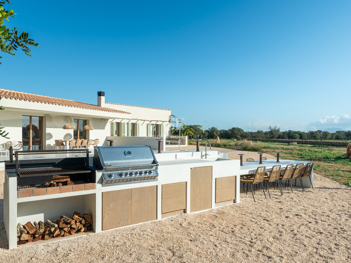 Modern outdoor kitchen with stainless steel grill, wood storage, and large dining table on a gravel terrace.