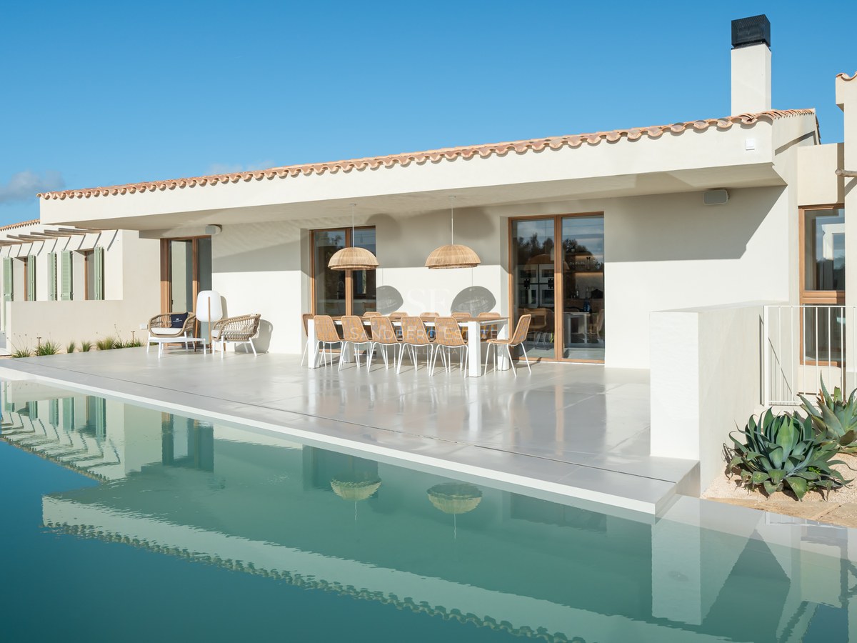 Modern white villa terrace with a large infinity pool, outdoor dining area, and clear blue sky background.