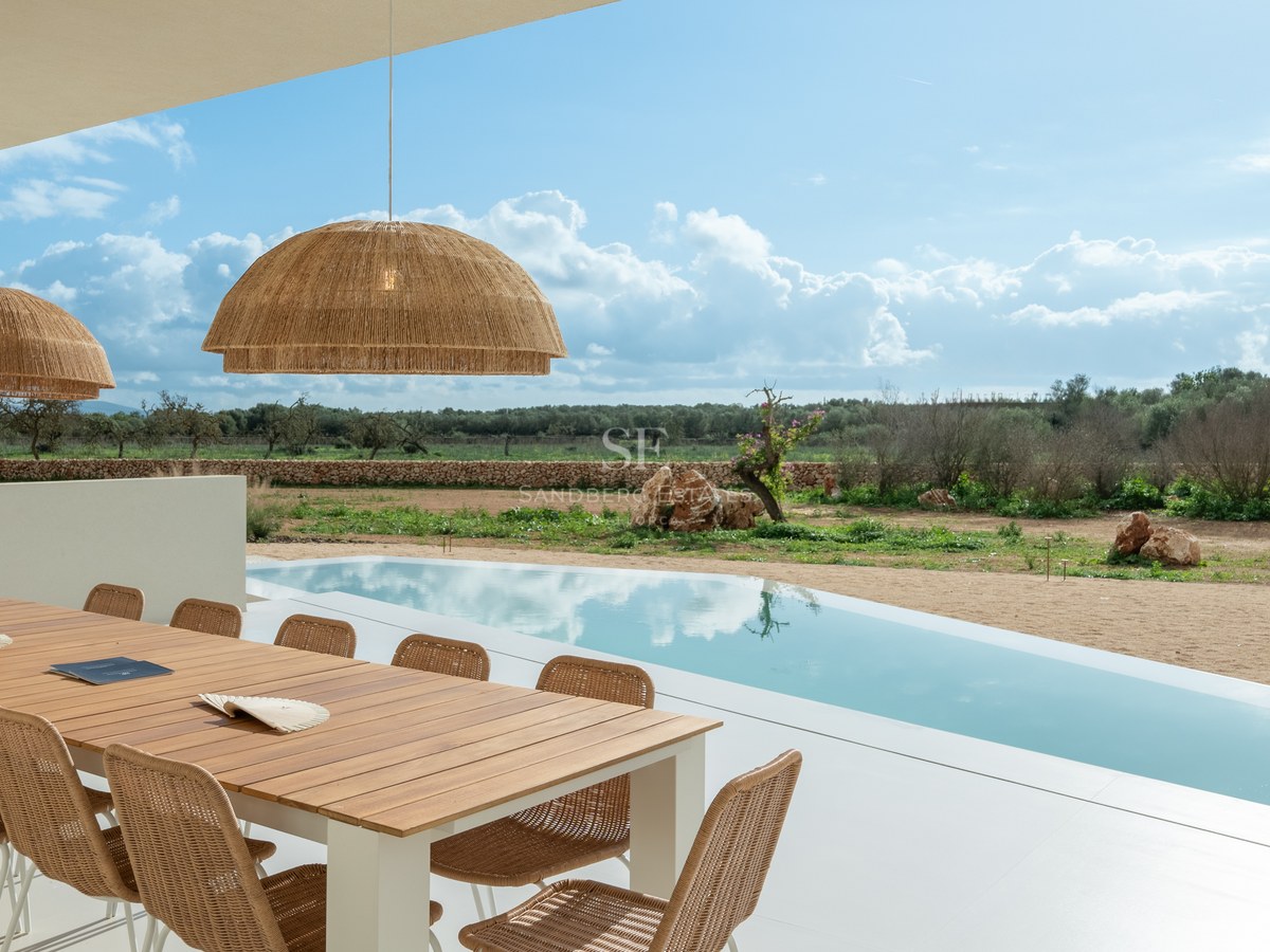 Modern wooden dining table and wicker chairs under a covered terrace next to a pool and rural landscape.