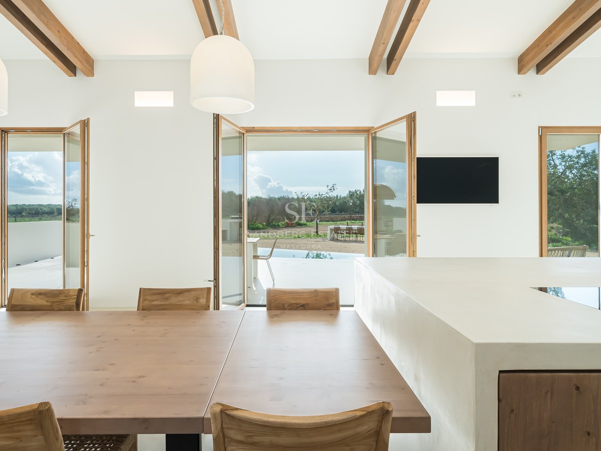 Modern kitchen with a large white island and wooden dining table featuring exposed ceiling beams and terrace views.