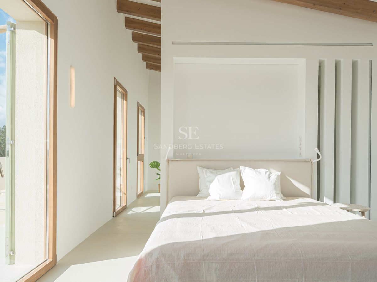 Bright master bedroom featuring exposed wooden ceiling beams, micro-cement floors, and floor-to-ceiling wooden glass doors.