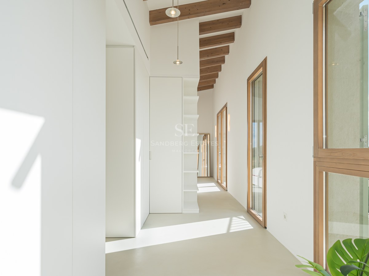 Bright white hallway featuring built-in wardrobes, exposed wooden ceiling beams, and large wood-framed windows.