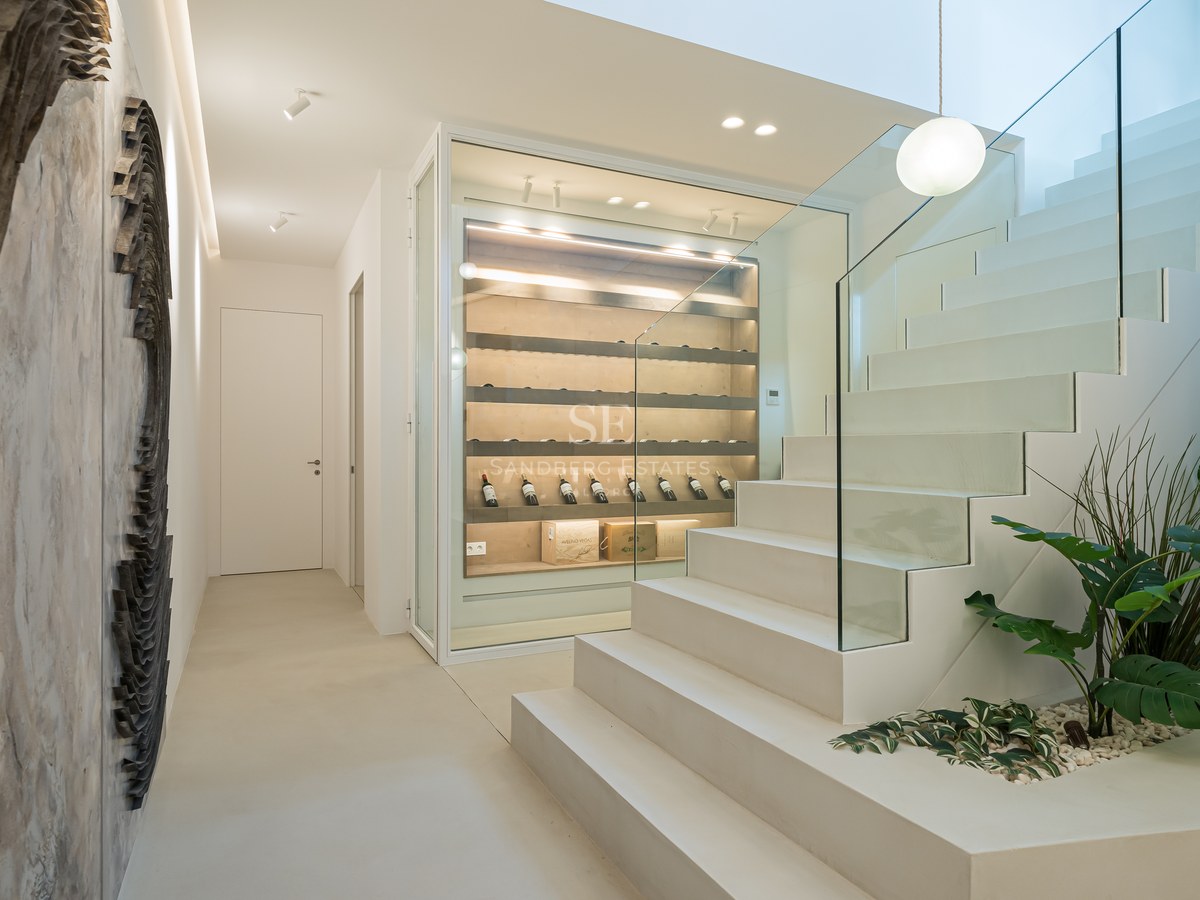 Modern white staircase with glass railing next to a glass-enclosed illuminated wine display case.