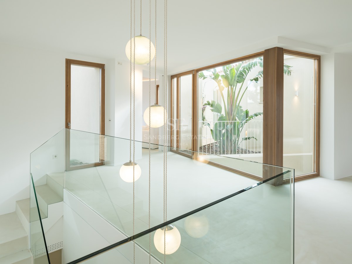 Interior view of a modern hallway with glass railings, wooden window frames, and hanging globe lights over a patio.