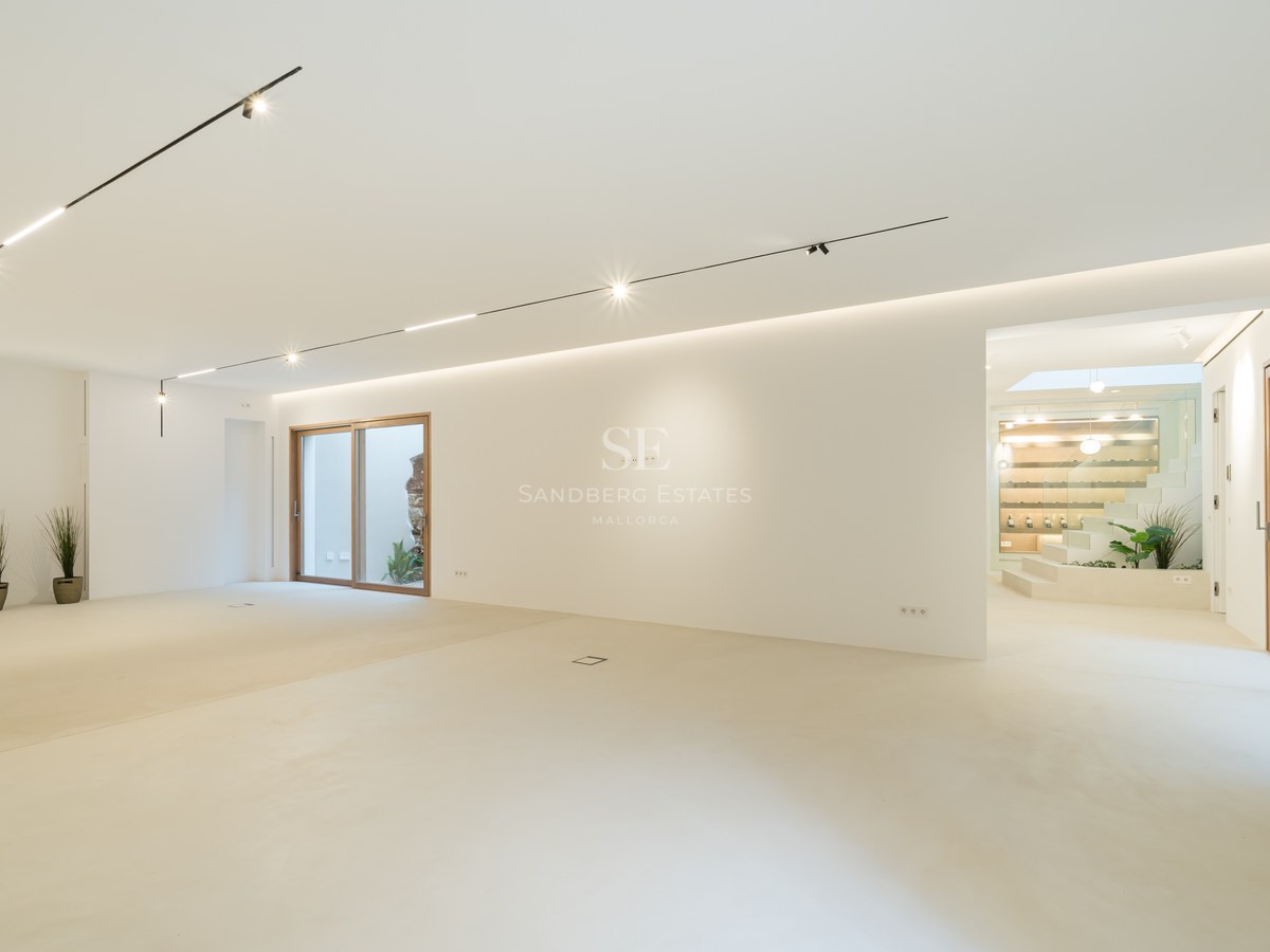 Large empty open-plan room with white walls, polished cream floors, track lighting, and wood-framed glass doors.