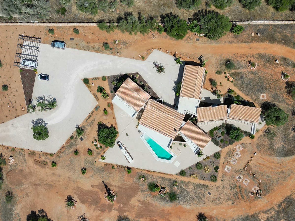 High-angle drone shot of a white modern villa with terracotta roofs, a geometric pool, and red soil grounds.