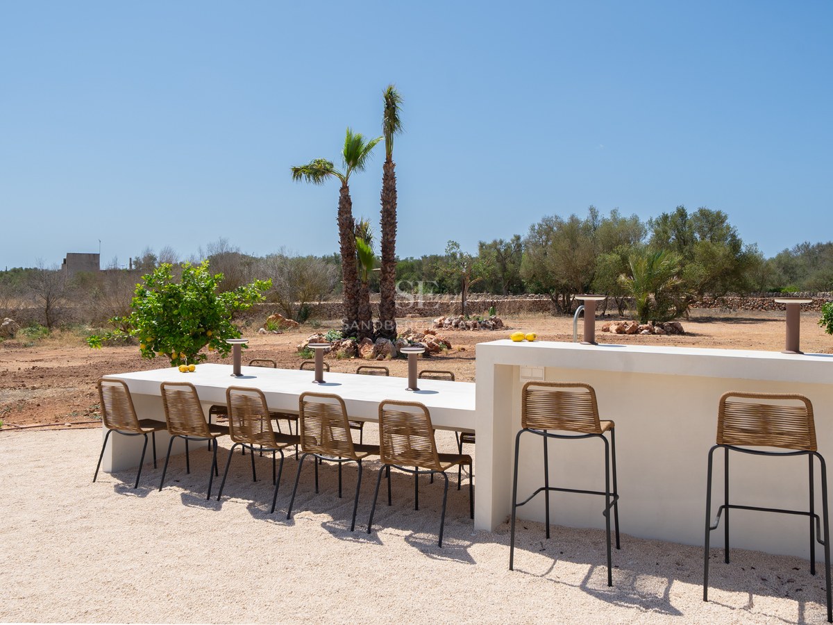 A long white dining table and bar counter with woven chairs on a gravel terrace under a clear blue sky.