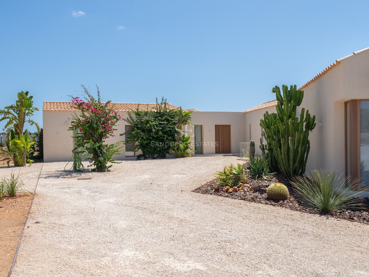 Contemporary cream-colored villa exterior with gravel courtyard, wooden doors, and desert-style landscaping under a clear sky.