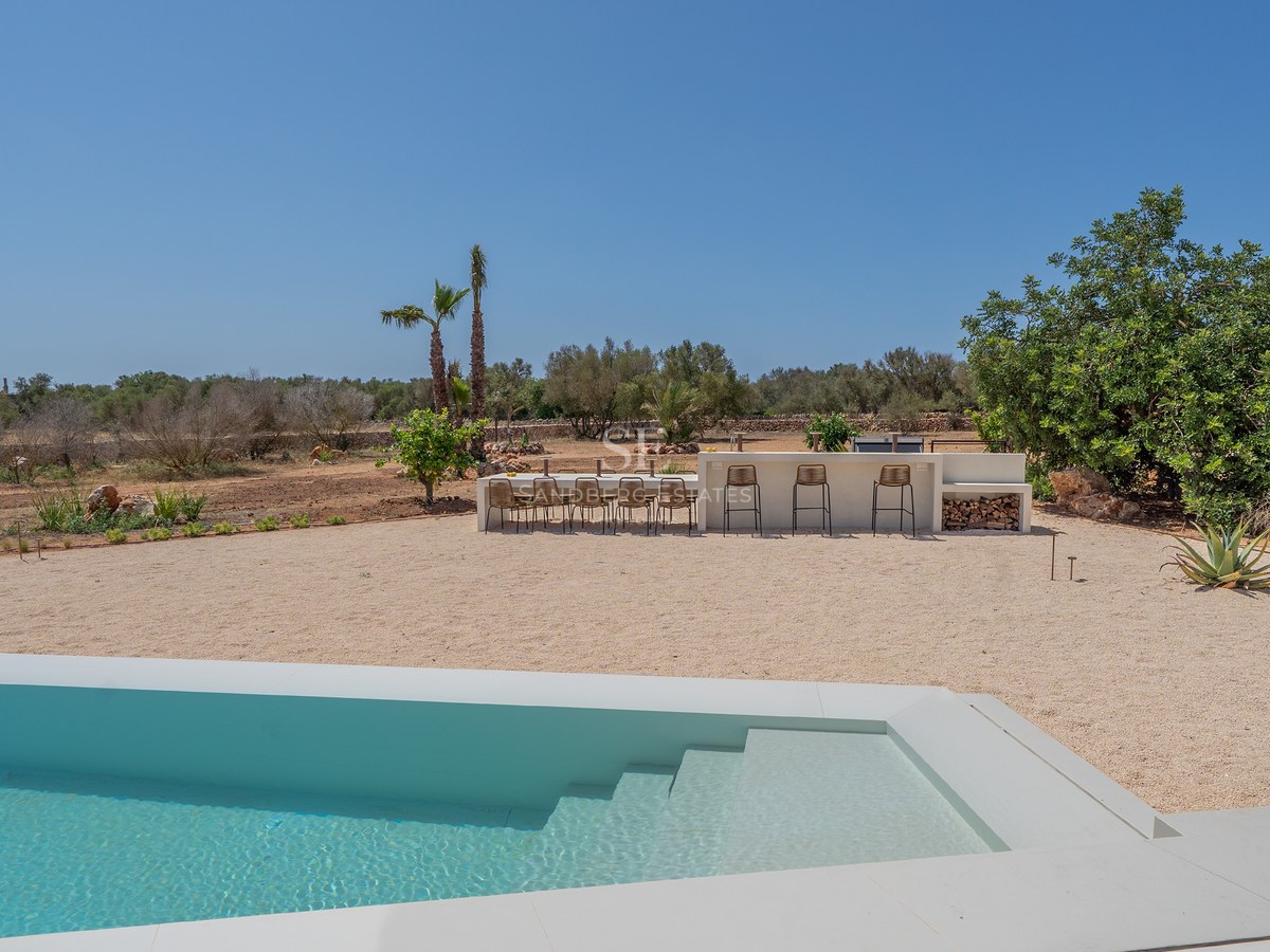 Modern white swimming pool foreground with outdoor dining area and bar set against a Mediterranean landscape.