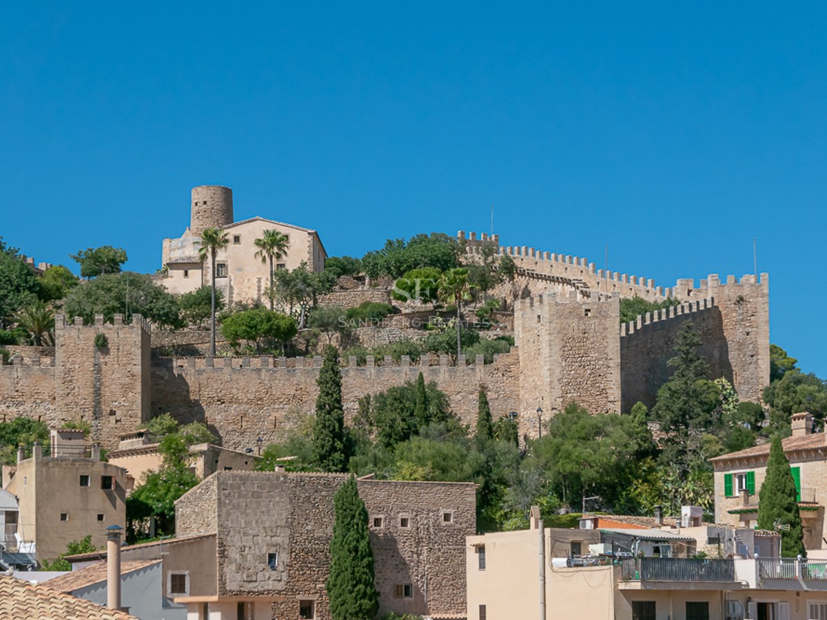 Steinmauern und Türme einer mittelalterlichen Festung auf einem Hügel unter blauem Himmel in einem mallorquinischen Dorf.
