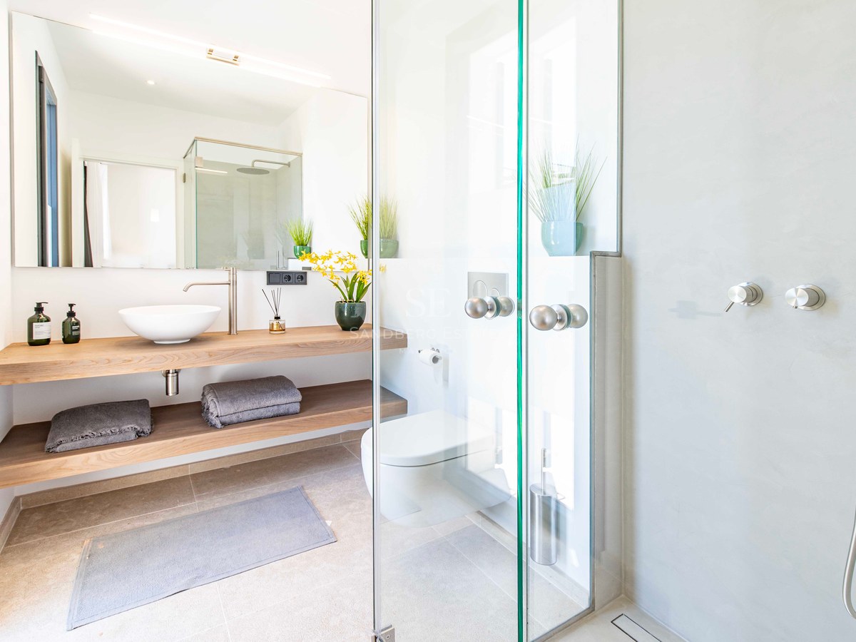 Salle de bains lumineuse avec douche à l'italienne, étagères en chêne, vasque blanche et toilettes suspendues.