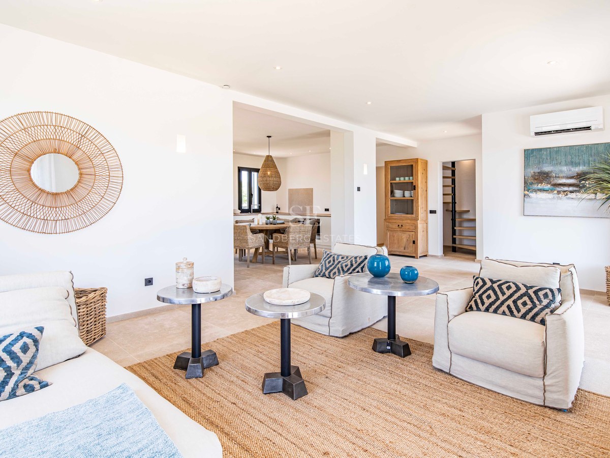 Salon lumineux avec murs blancs, sol en pierre, tapis en jute, fauteuils beiges et tables d'appoint en métal.