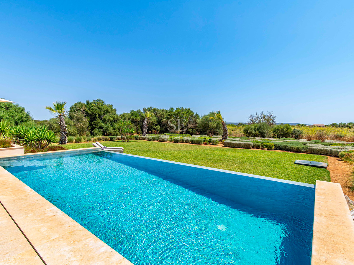 Piscine à débordement aux eaux turquoise bordée d'une pelouse verdoyante et d'arbres méditerranéens sous un ciel bleu.