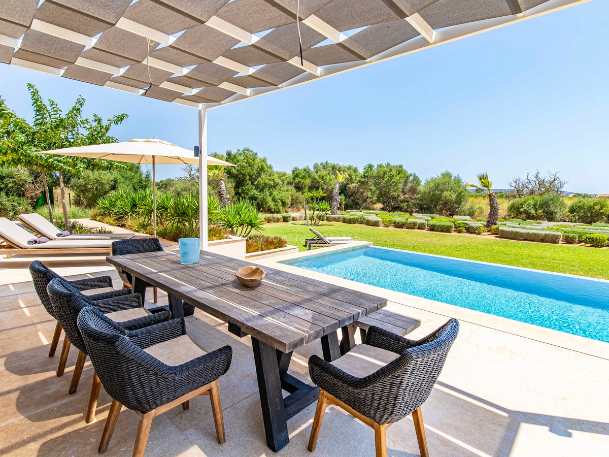 Espace repas extérieur avec table en bois et chaises en osier sous un voile d'ombrage, face à une piscine et un jardin.