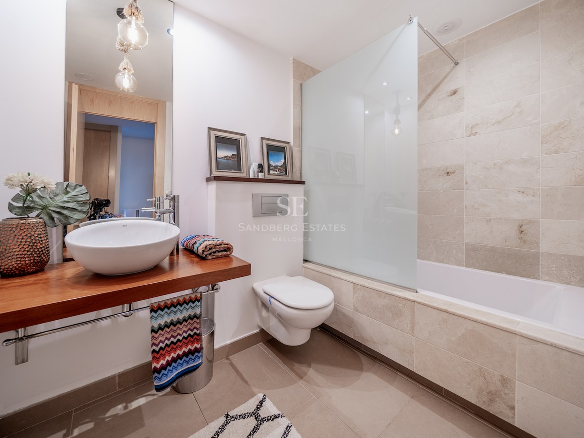 Modern bathroom featuring a wooden vanity, vessel sink, wall-hung toilet, and a bathtub with stone tile walls.