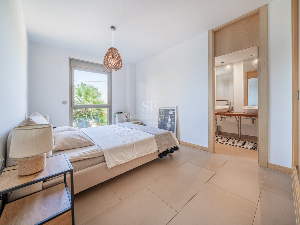 Bright modern bedroom with light wood accents, an ensuite bathroom with double sinks, and a window showing palm trees.