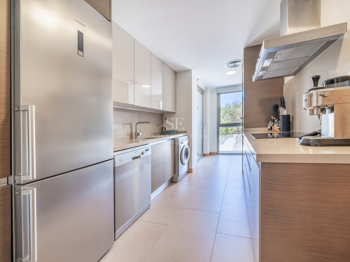 Galley-style kitchen with light wood cabinetry, stainless steel appliances, and large window at the end.