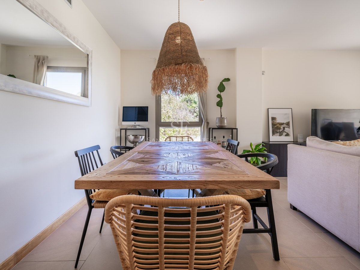 A large wooden parquetry dining table with a rattan pendant light in a bright, modern open-plan living room.