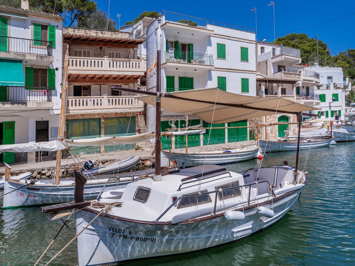 Traditional white houses with green shutters overlooking a calm harbor with classic wooden boats on turquoise water.