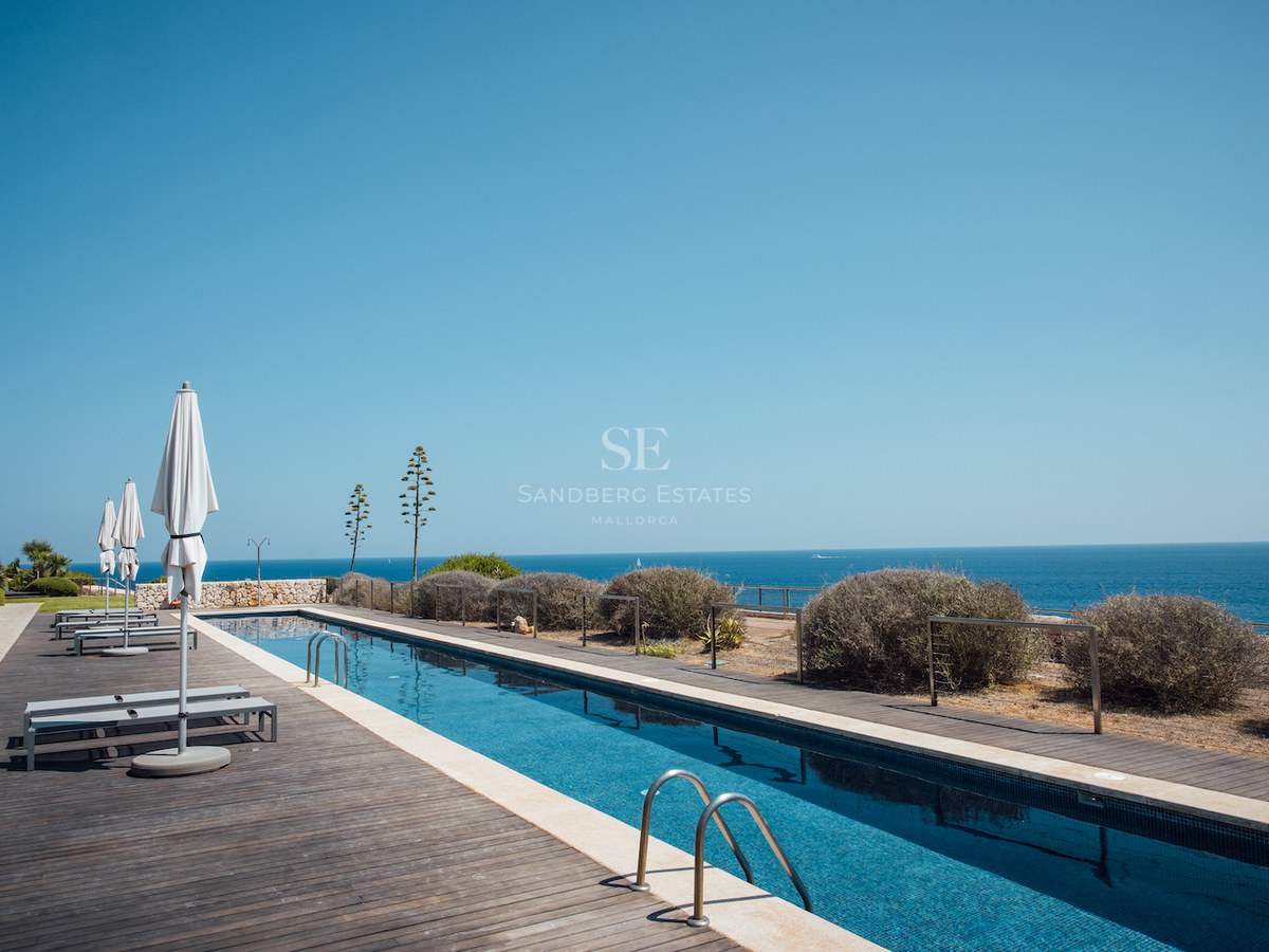 A long rectangular swimming pool with a dark wooden deck, white umbrellas, and an expansive view of the blue sea.
