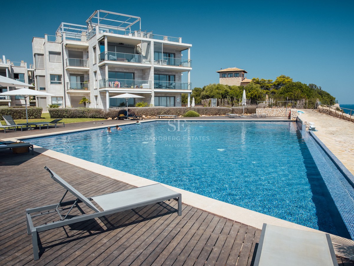 Large outdoor swimming pool with wooden deck, sun loungers, and modern apartment building under a clear blue sky.