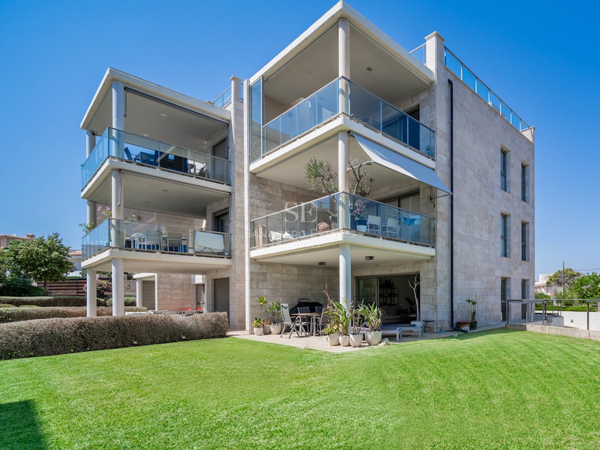Three-story contemporary villa featuring natural stone cladding, glass balconies, and a lush lawn under a clear blue sky.