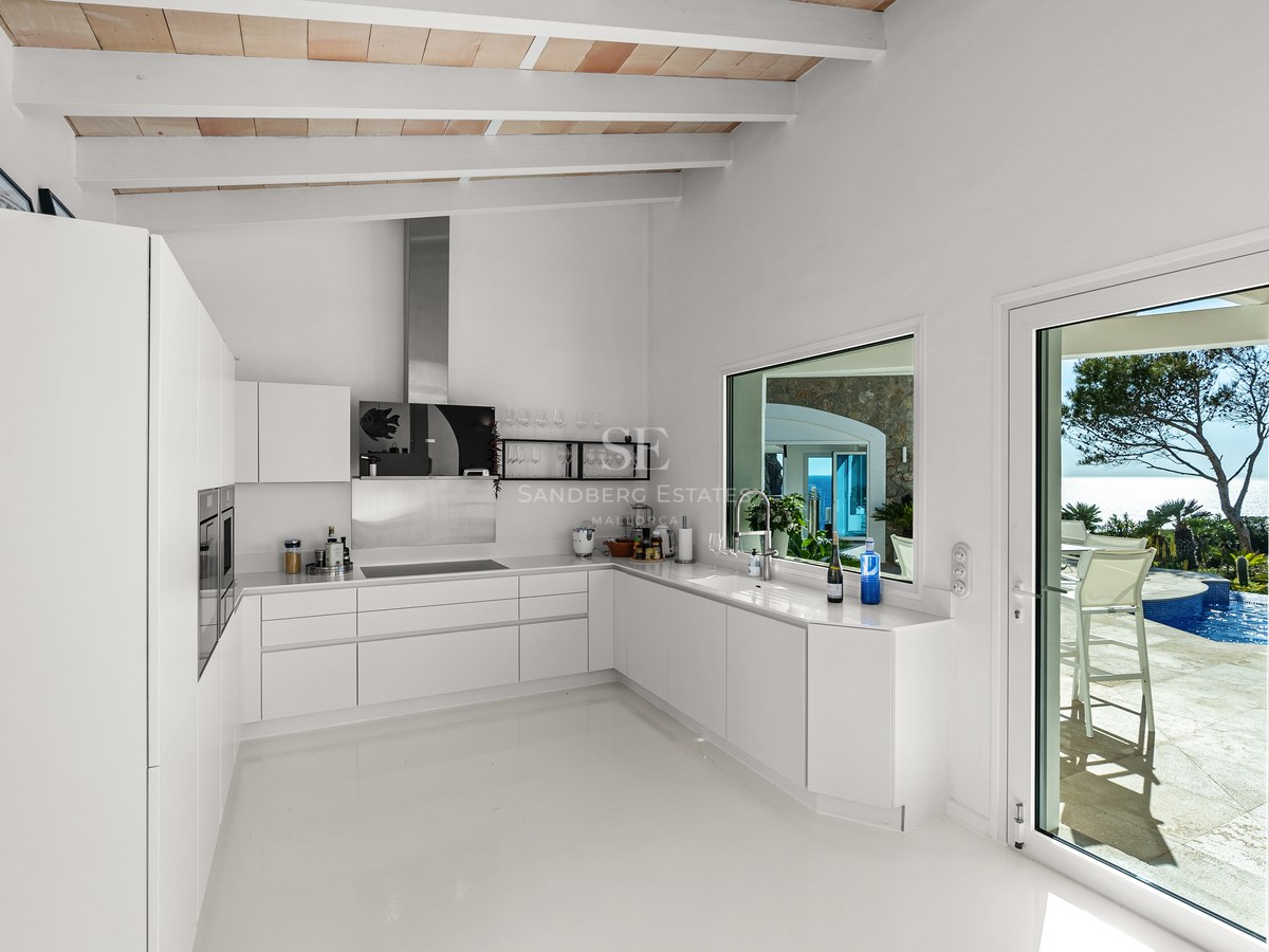 Sleek white minimalist kitchen featuring exposed wooden beams and glass door leading to a pool terrace with sea views.