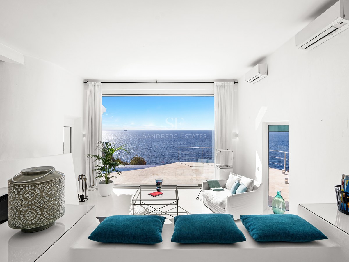 Bright white minimalist living room with floor-to-ceiling windows overlooking a Mediterranean terrace and the blue sea.