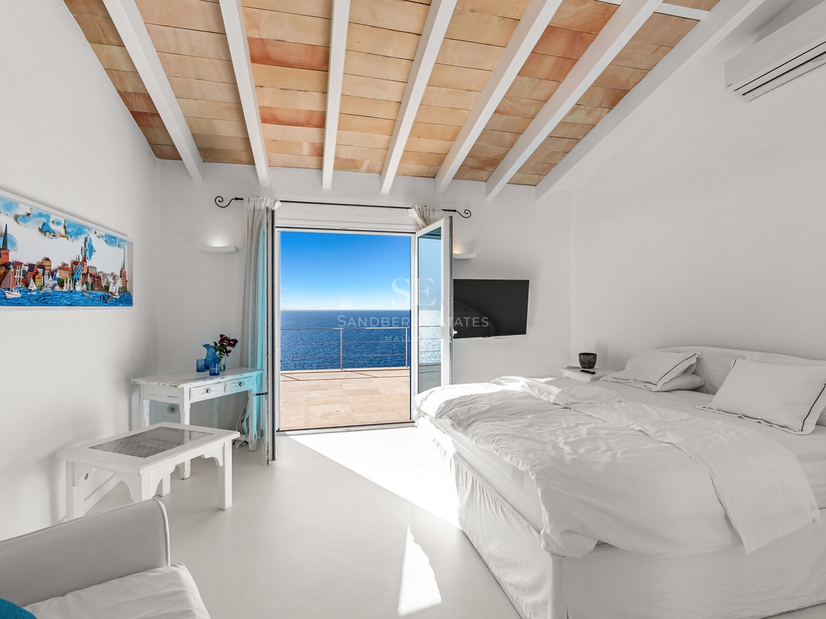 Bright bedroom with white linens, terracotta-tiled ceiling, and open doors leading to a terrace overlooking the blue sea.