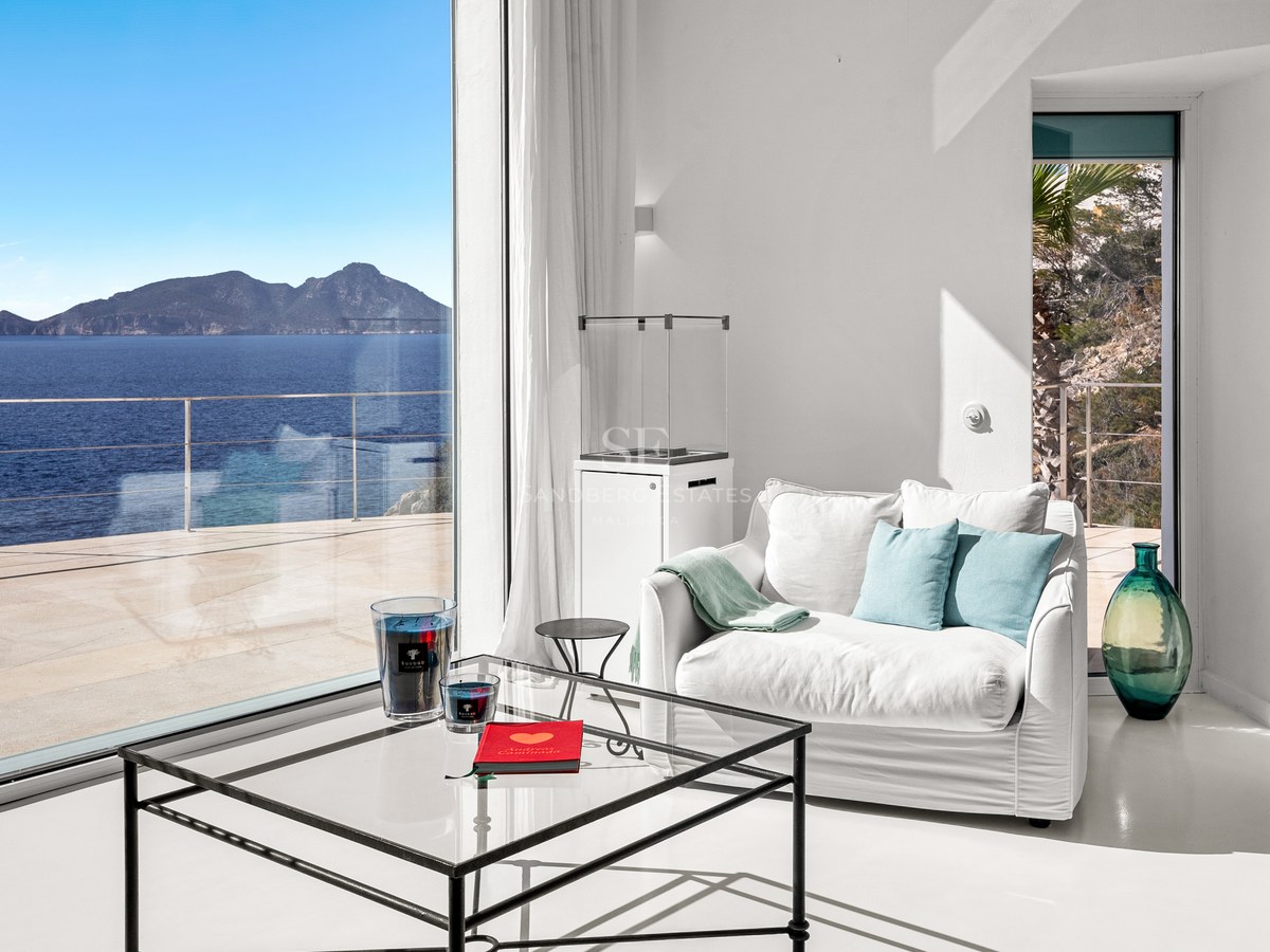 Interior of a white minimalist living room featuring a glass coffee table and armchair overlooking the sea.