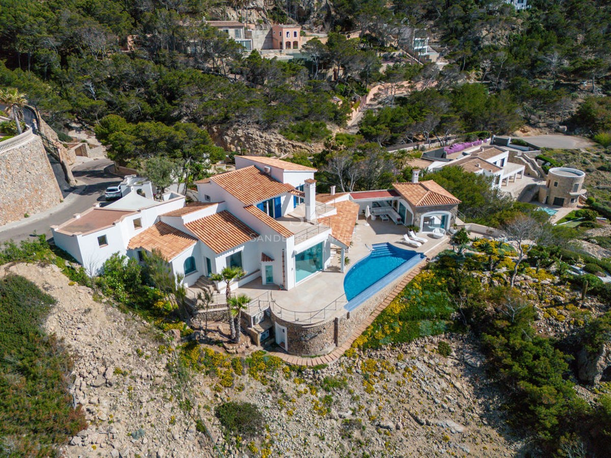 Aerial view of a white multi-level villa with terracotta roofs, infinity pool, and stone walls on a lush hillside.