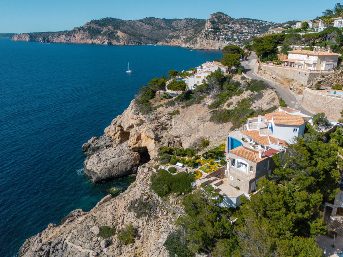 Drone view of a Mediterranean villa perched on a cliff above blue sea with a rocky coastline.