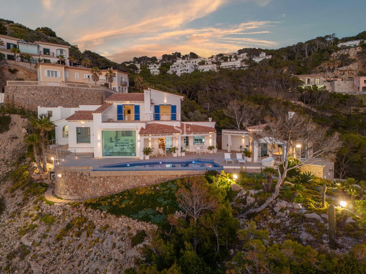Evening view of a white multi-level villa on a rocky cliff with a curved pool, terracotta roof, and local flora.