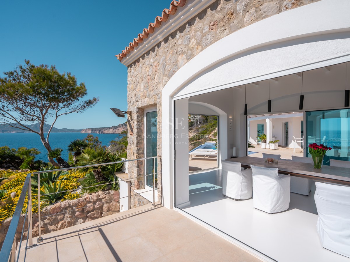 Modern dining room with wood table and white chairs opening to a sunny terrace with panoramic Mediterranean sea views.