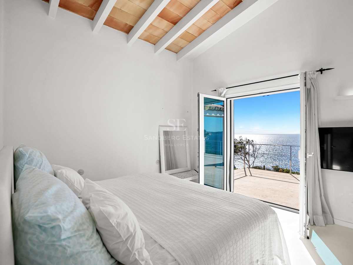 Bright master bedroom featuring white exposed ceiling beams and glass doors opening to a panoramic Mediterranean sea view.