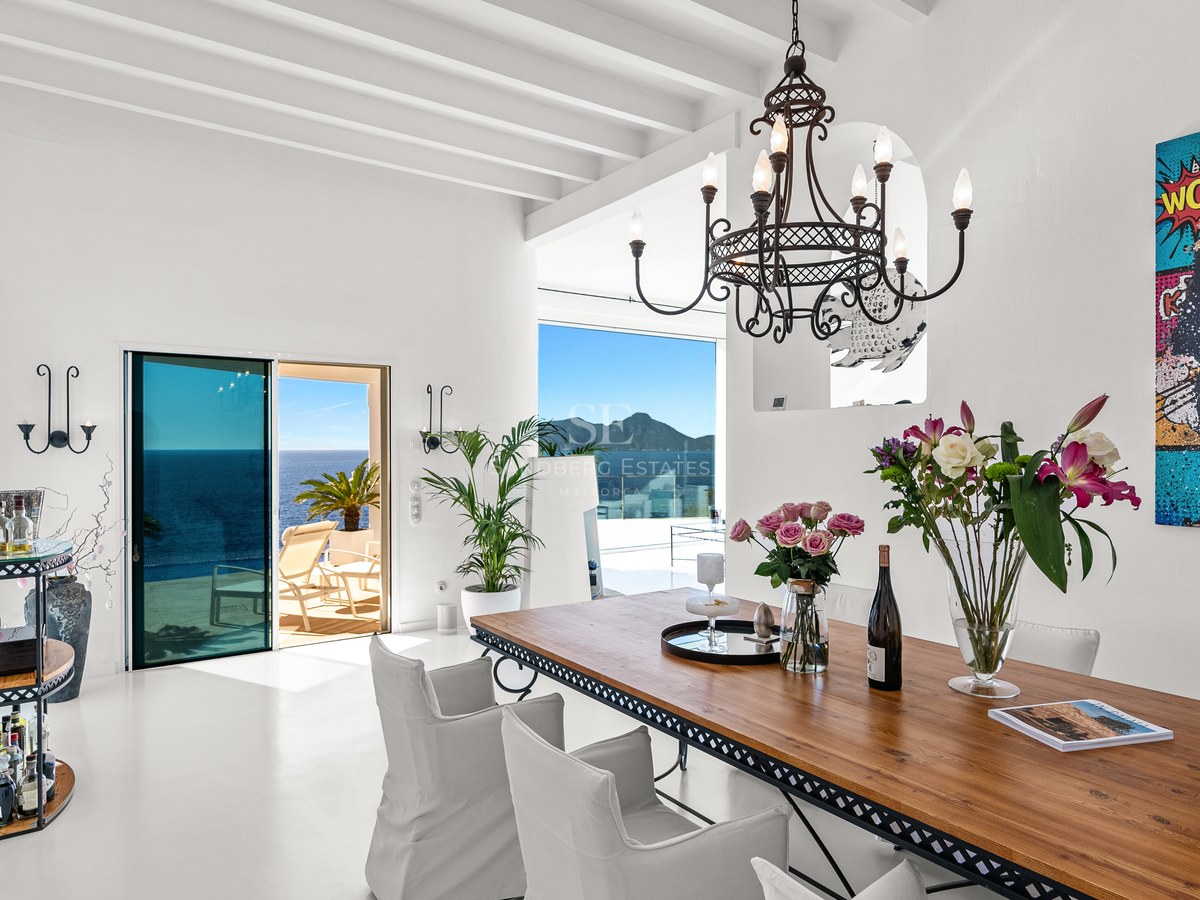 Modern dining room with a large wooden table, white beamed ceiling, and floor-to-ceiling windows overlooking the Mediterranean.