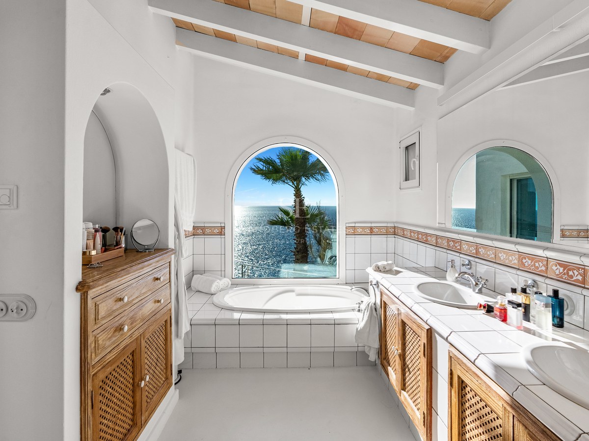 Bright white bathroom featuring a bathtub set before an arched window with Mediterranean sea views and beamed ceiling.