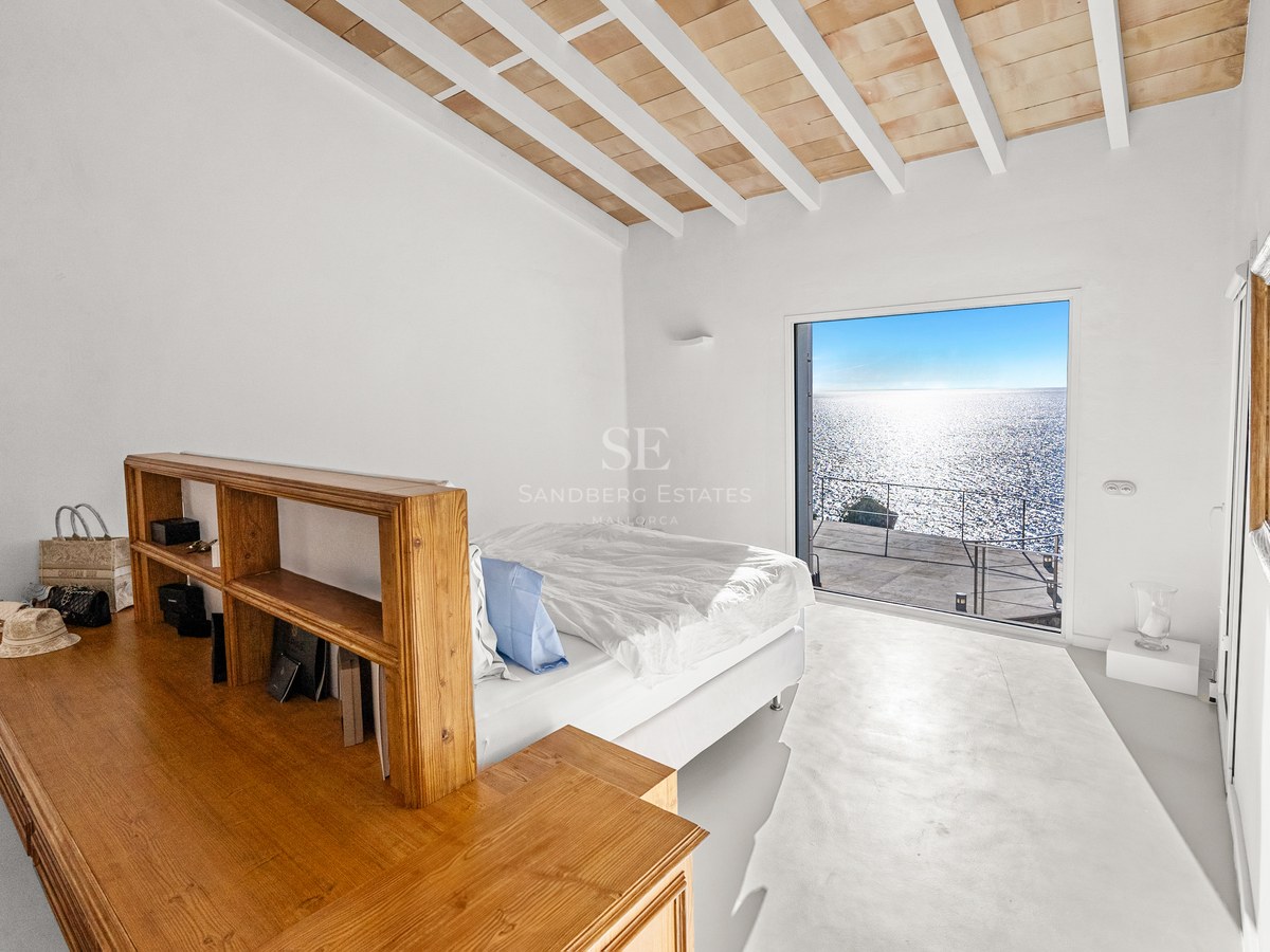 Minimalist white bedroom with custom wooden furniture and a large window overlooking the sparkling Mediterranean sea.