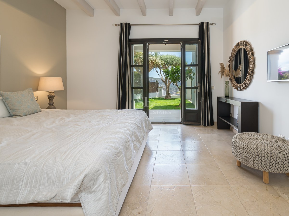 Chambre spacieuse avec sol en pierre naturelle, poutres blanches et portes vitrées ouvrant sur un jardin verdoyant.
