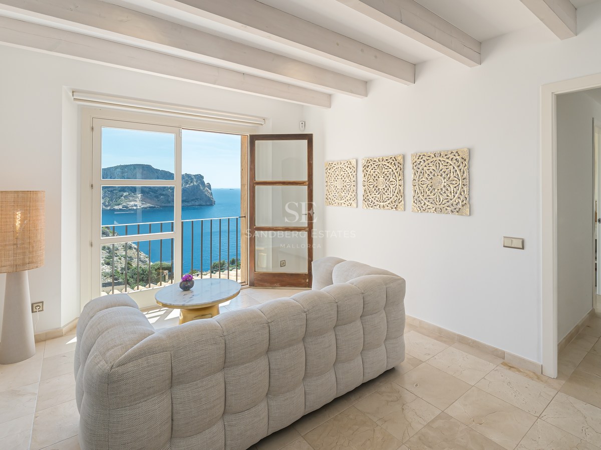 Salon lumineux avec poutres blanches, sol en pierre naturelle et balcon donnant sur la mer bleue et les falaises.