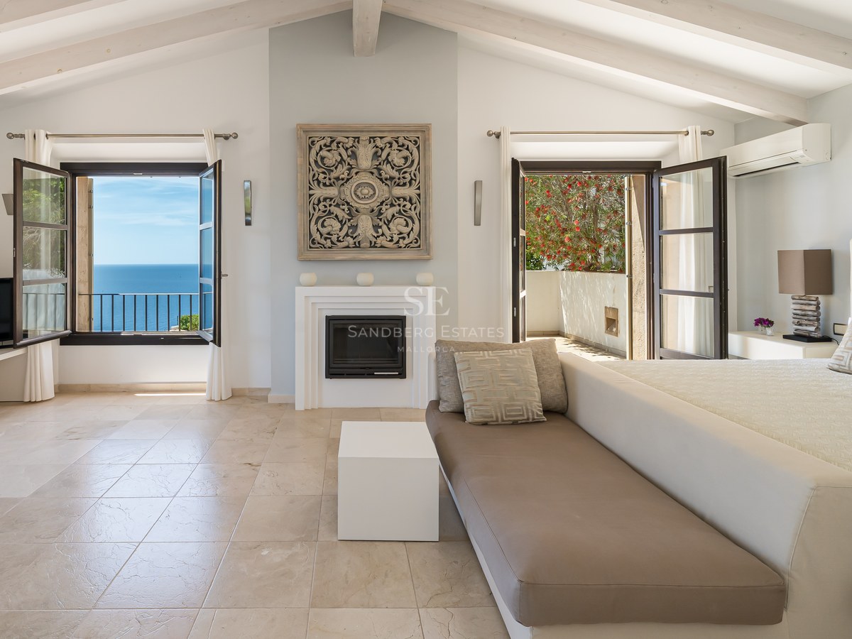 Grande chambre avec plafonds à poutres blanches, cheminée, sol en pierre et fenêtres ouvertes sur la mer.