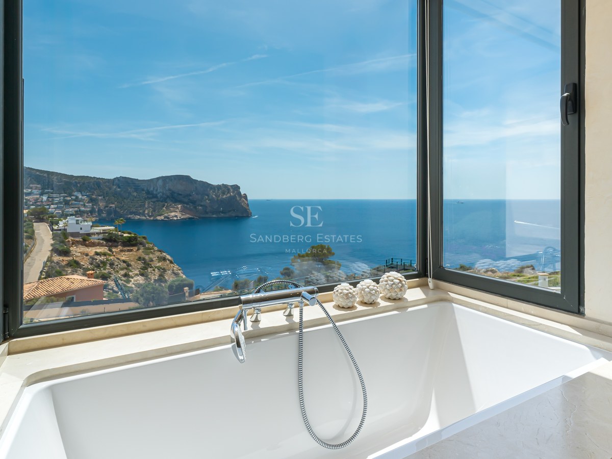 Baignoire blanche moderne devant de grandes baies vitrées donnant sur une côte rocheuse et la mer bleue.