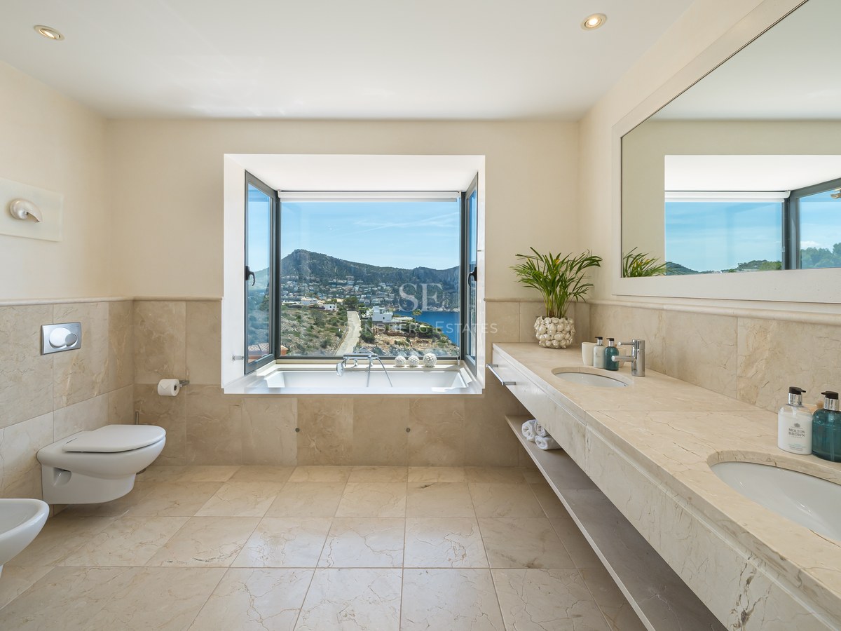 Salle de bain en travertin beige avec double vasque et baignoire face à une fenêtre ouvrant sur la mer et les montagnes.