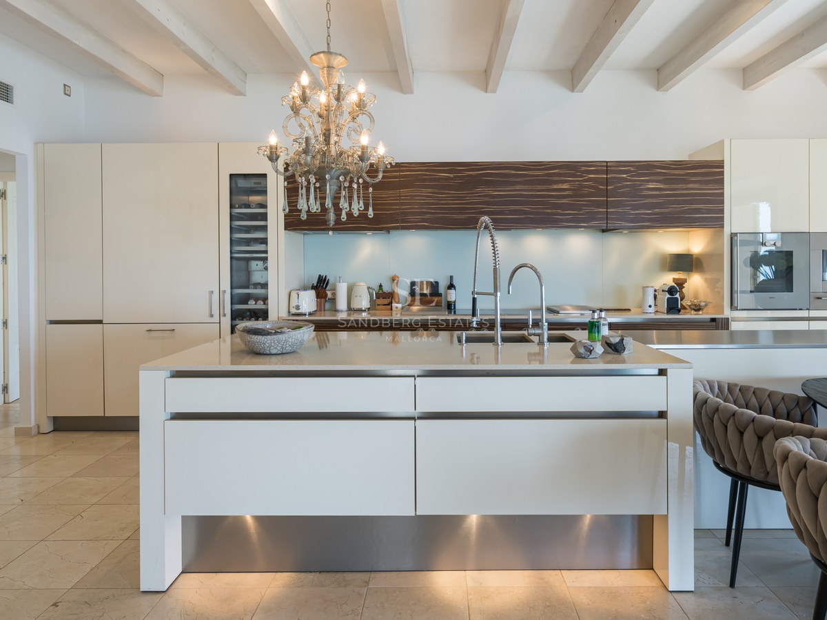 Cuisine moderne blanche avec grand îlot, lustre en cristal et plafonds à poutres apparentes.
