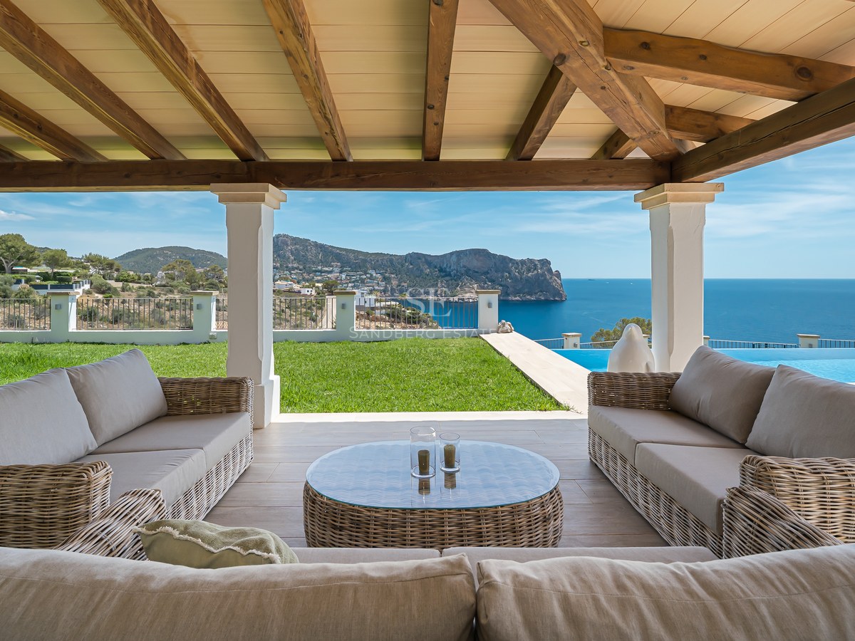 Terrasse couverte avec canapés en osier et poutres en bois donnant sur un jardin, une piscine et la mer.