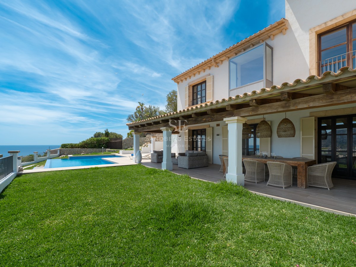 Façade de villa blanche avec terrasse couverte, pelouse et piscine surplombant la mer sous un ciel bleu.