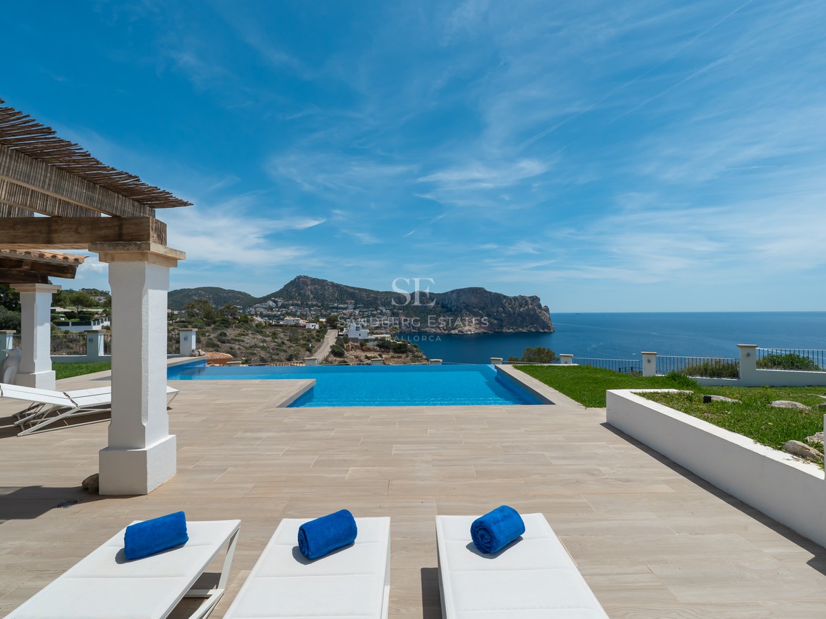 Terrasse de luxe avec piscine à débordement, bains de soleil blancs et pergola en bois surplombant la côte.