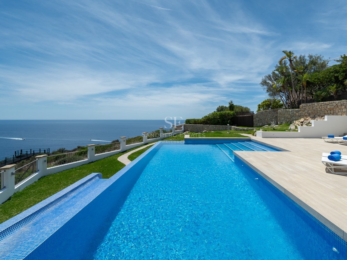 Piscine à débordement bleue surplombant la mer avec transats sur une terrasse en pierre sous un ciel bleu.